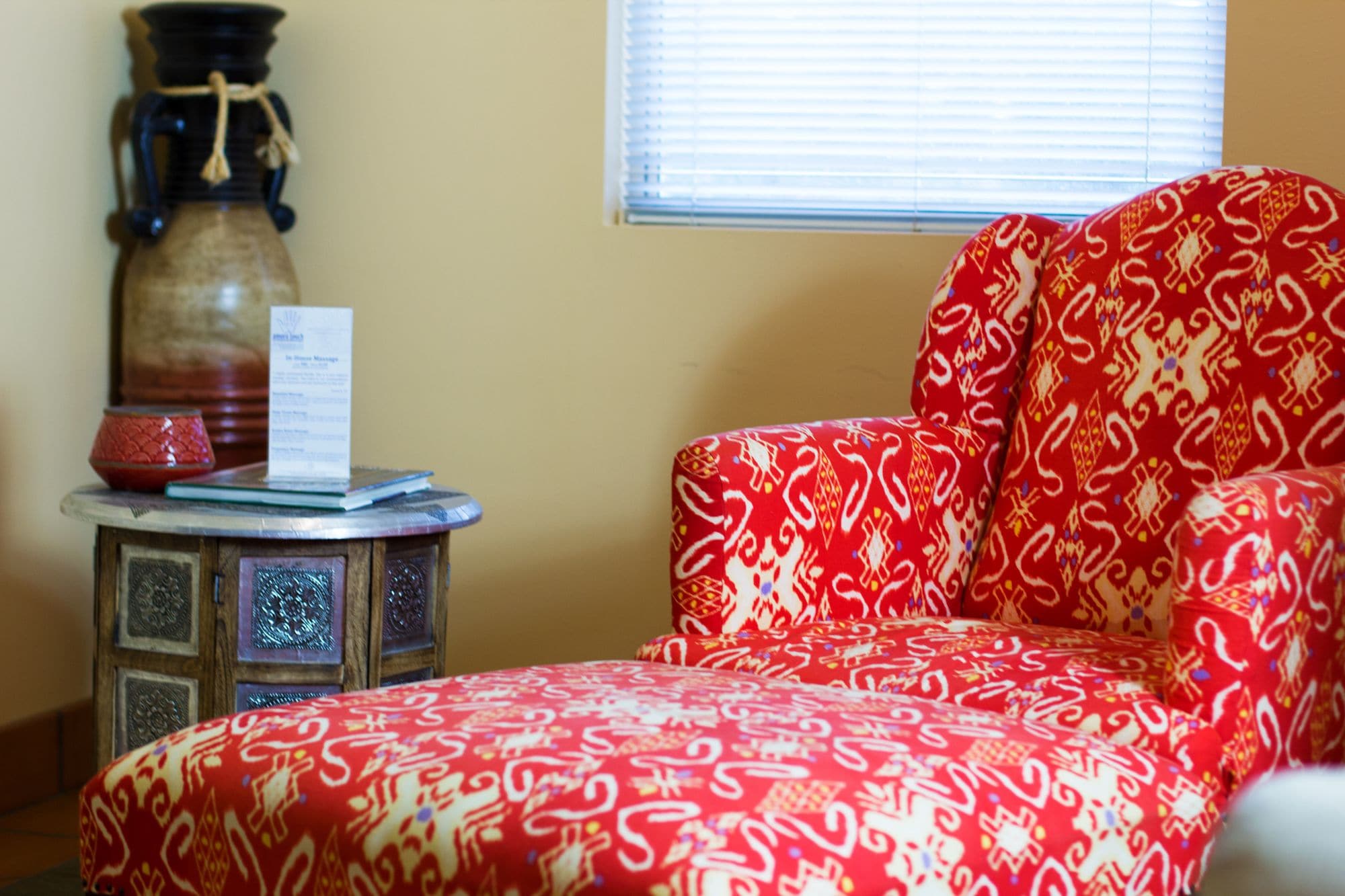 A room with a patterned red sofa, a small side table with a vase and a menu, and a window with blinds.