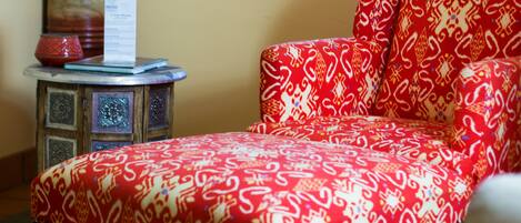 A room with a patterned red sofa, a small side table with a vase and a menu, and a window with blinds.