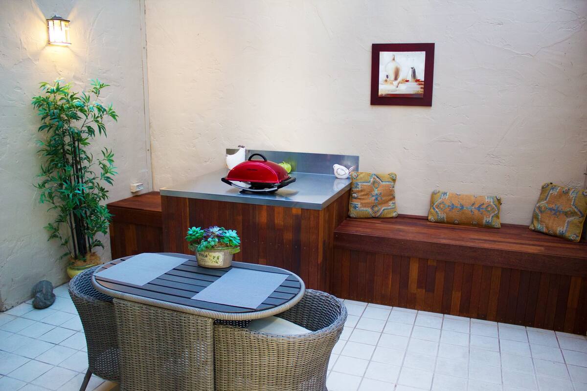 Un petit espace extérieur aménagé pour les repas, avec une table et des chaises en osier, un barbecue rouge et un banc doté de coussins.