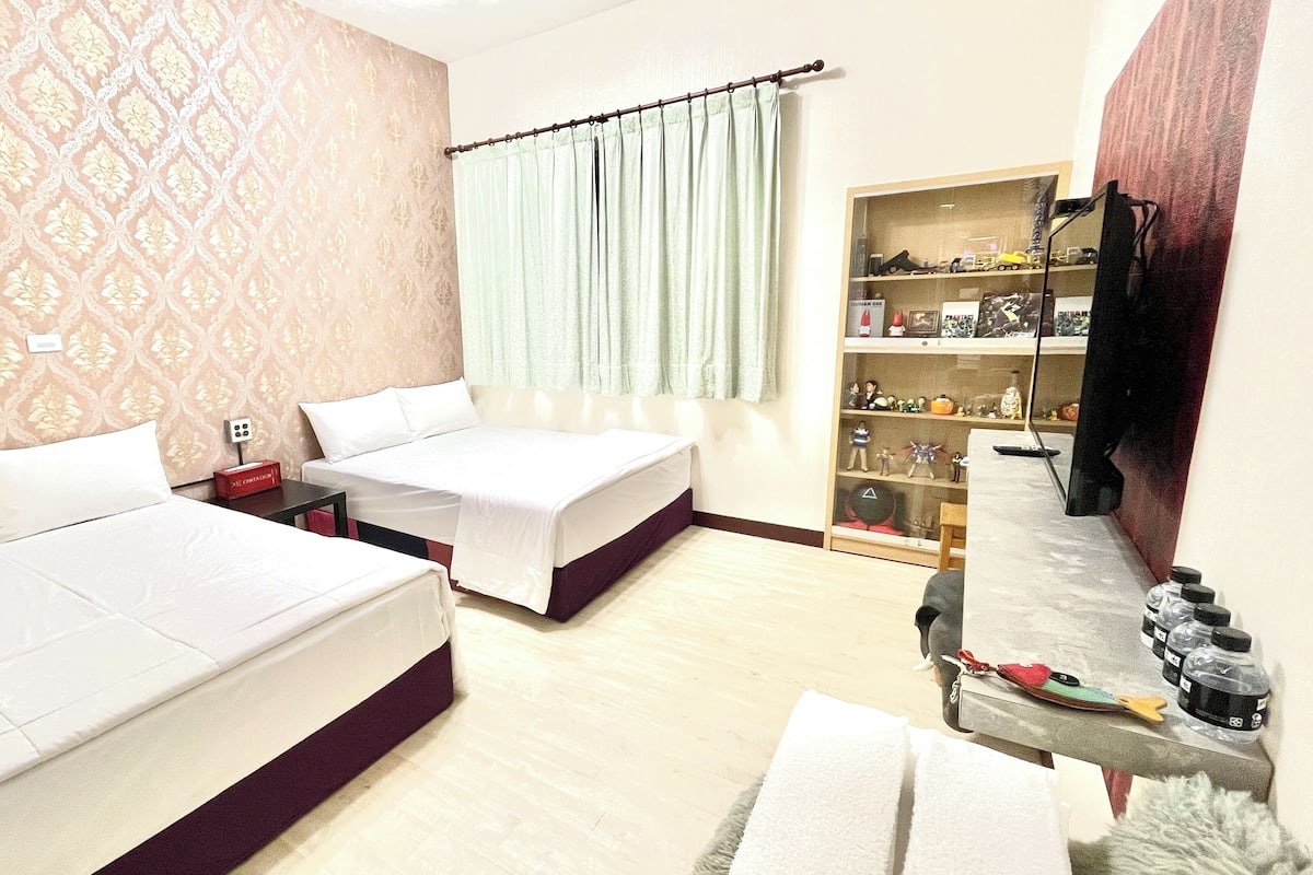 Quarto de hotel com uma cama grande, uma cama menor, uma escrivaninha e uma estante com diversos objetos.