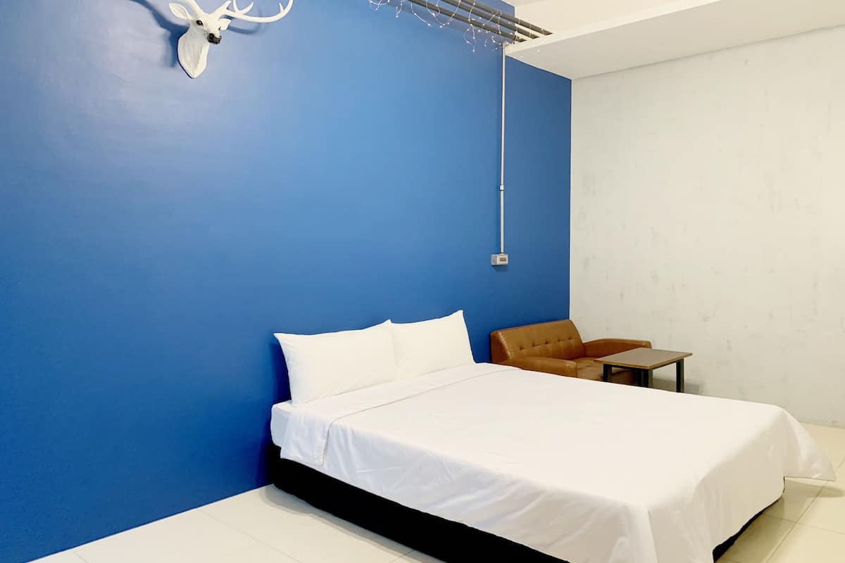 Quarto com cama, uma cadeira, uma mesinha e luminária fixada na parede.