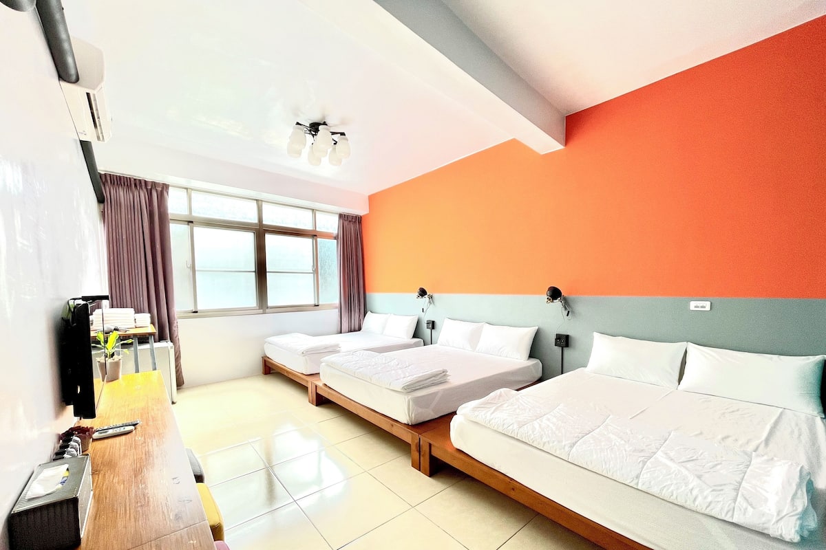 Quarto de hotel com duas camas, uma escrivaninha com computador, uma janela com cortinas e um lustre.