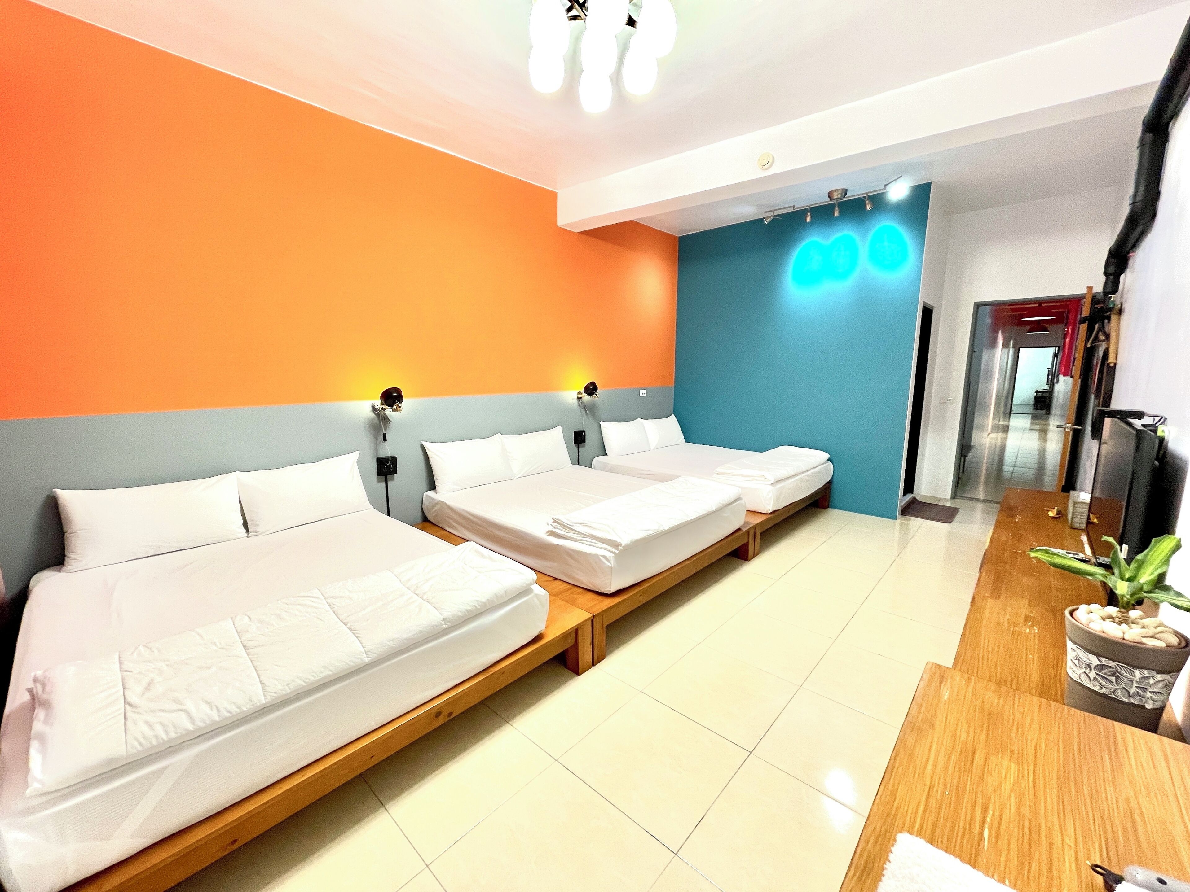 A hotel room with three single beds, a wooden bench, a desk with a plant, and a blue wall with a neon sign.
