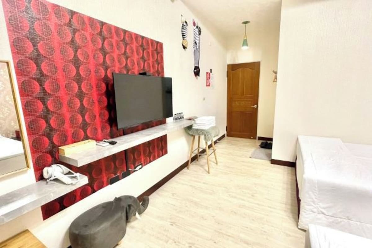 A hotel room with a TV on a red patterned wall, a wooden door, a white bed, and a small table with a chair.