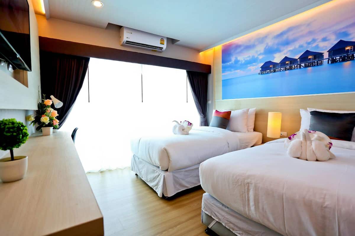 A hotel room with two beds, a desk, a potted plant, and a painting of overwater bungalows.