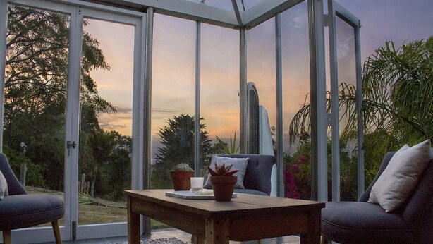 A conservatory with a glass door, a wooden table, two chairs, and a view of the sunset. - National Award Winner with Stunning View (Havelock North)