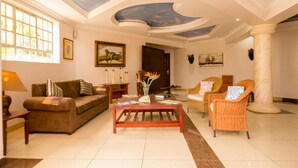 A spacious living room with a high ceiling, a large window, and a fireplace. - Hotel Rudi (Nairobi)