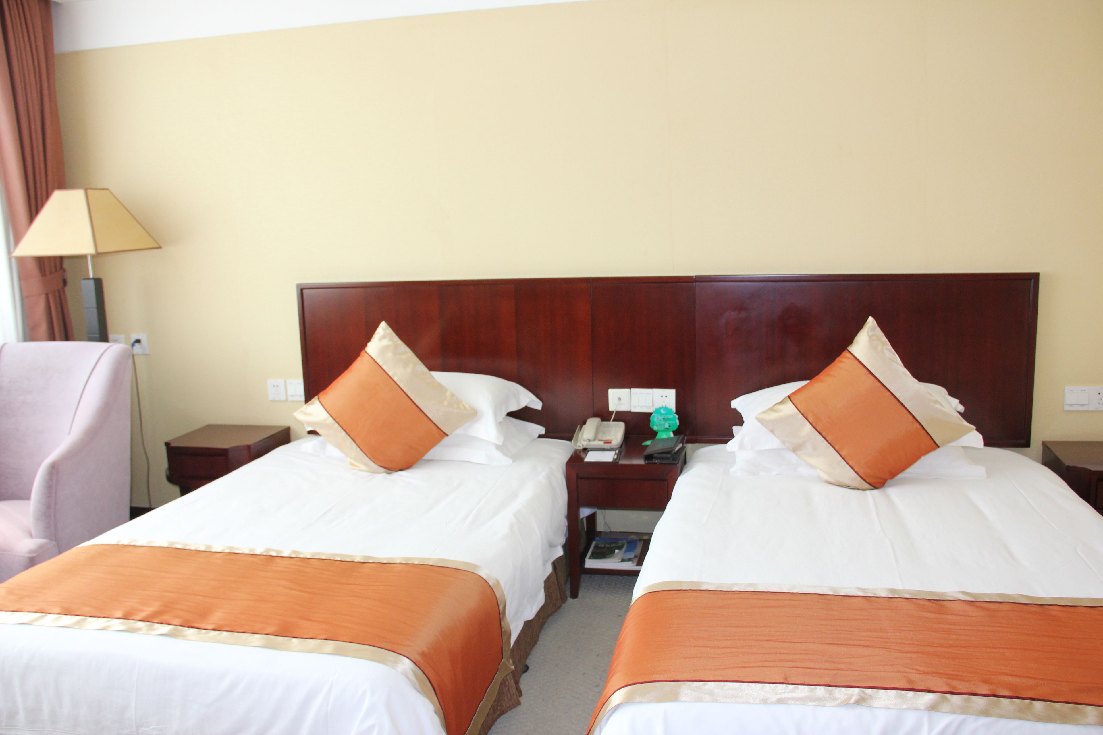 Une chambre d’hôtel avec deux lits, chacun doté d’une tête de lit en bois, de draps blancs et d’éléments décoratifs orange. Une table de chevet avec une lampe et un téléphone se trouve entre les deux lits.