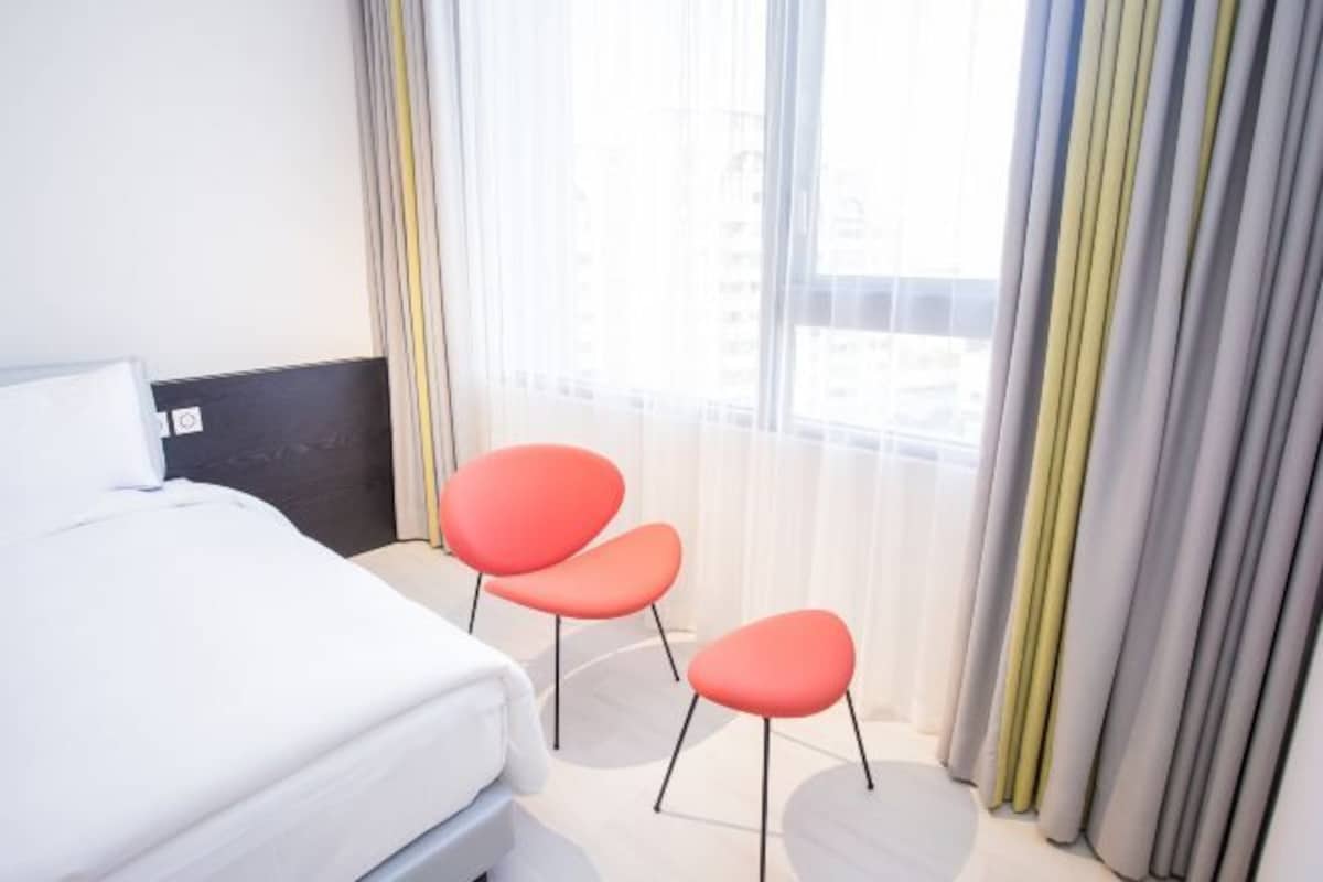 A hotel room with a bed, two red chairs, and a large window with sheer curtains.