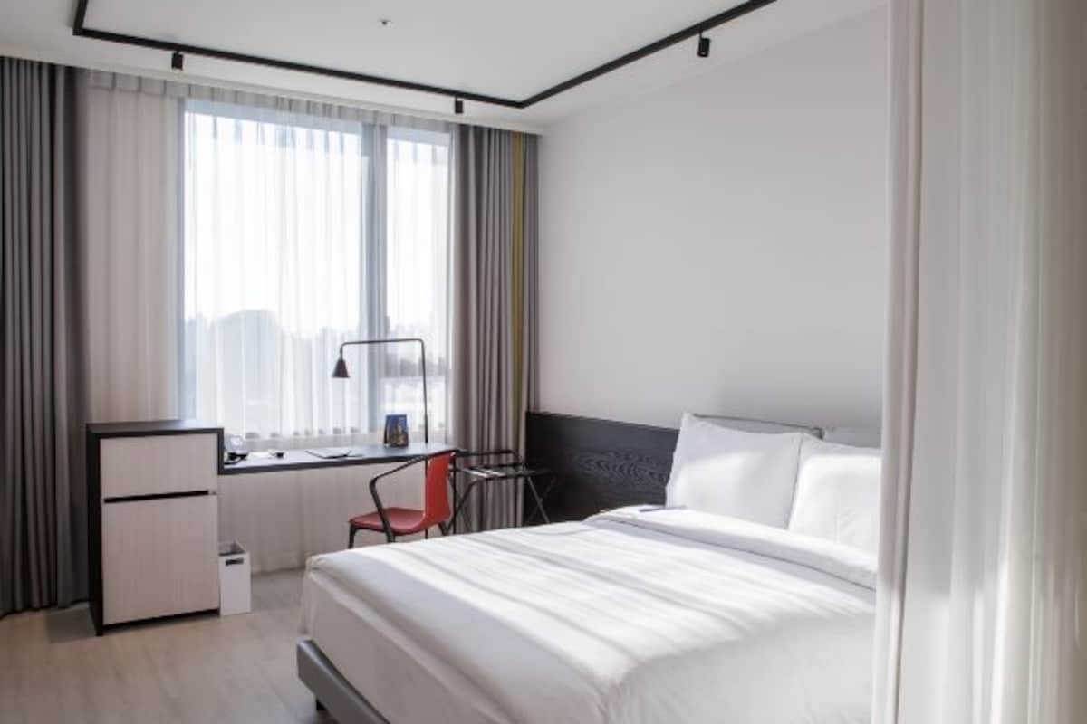 A modern hotel room with a bed, desk, chair, and a view of the city through large windows.