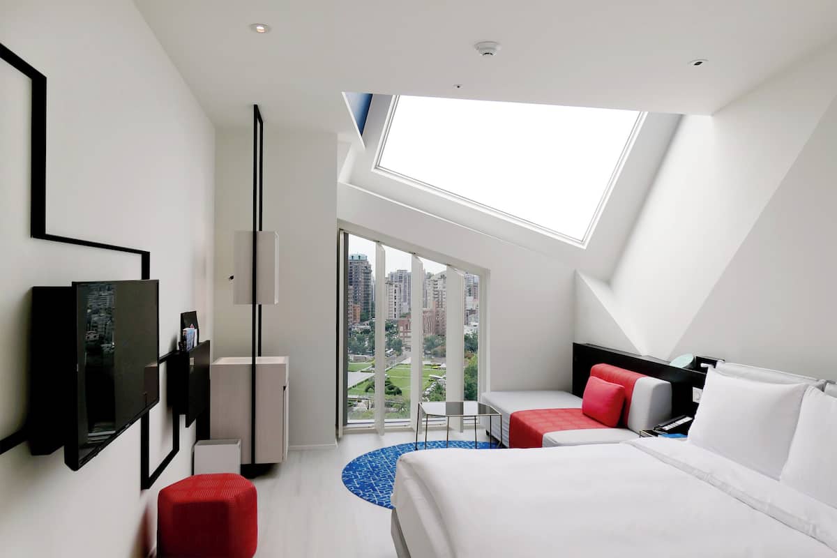 A modern hotel room with a large bed, a TV mounted on the wall, a small table, and a view of the city through large windows.