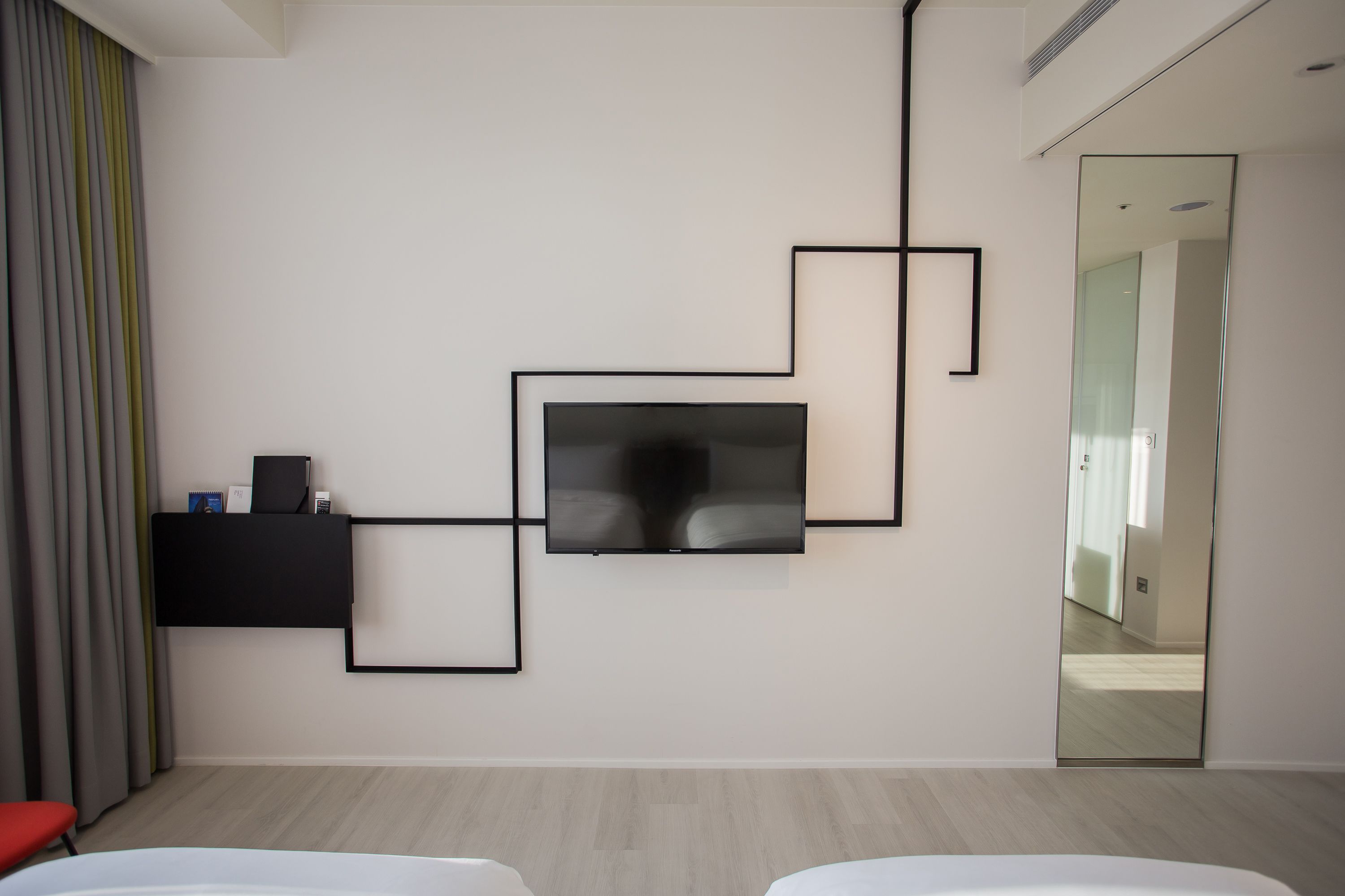 A modern hotel room with a flat-screen TV mounted on the wall, a small black shelf with items, and a large mirror reflecting the room.
