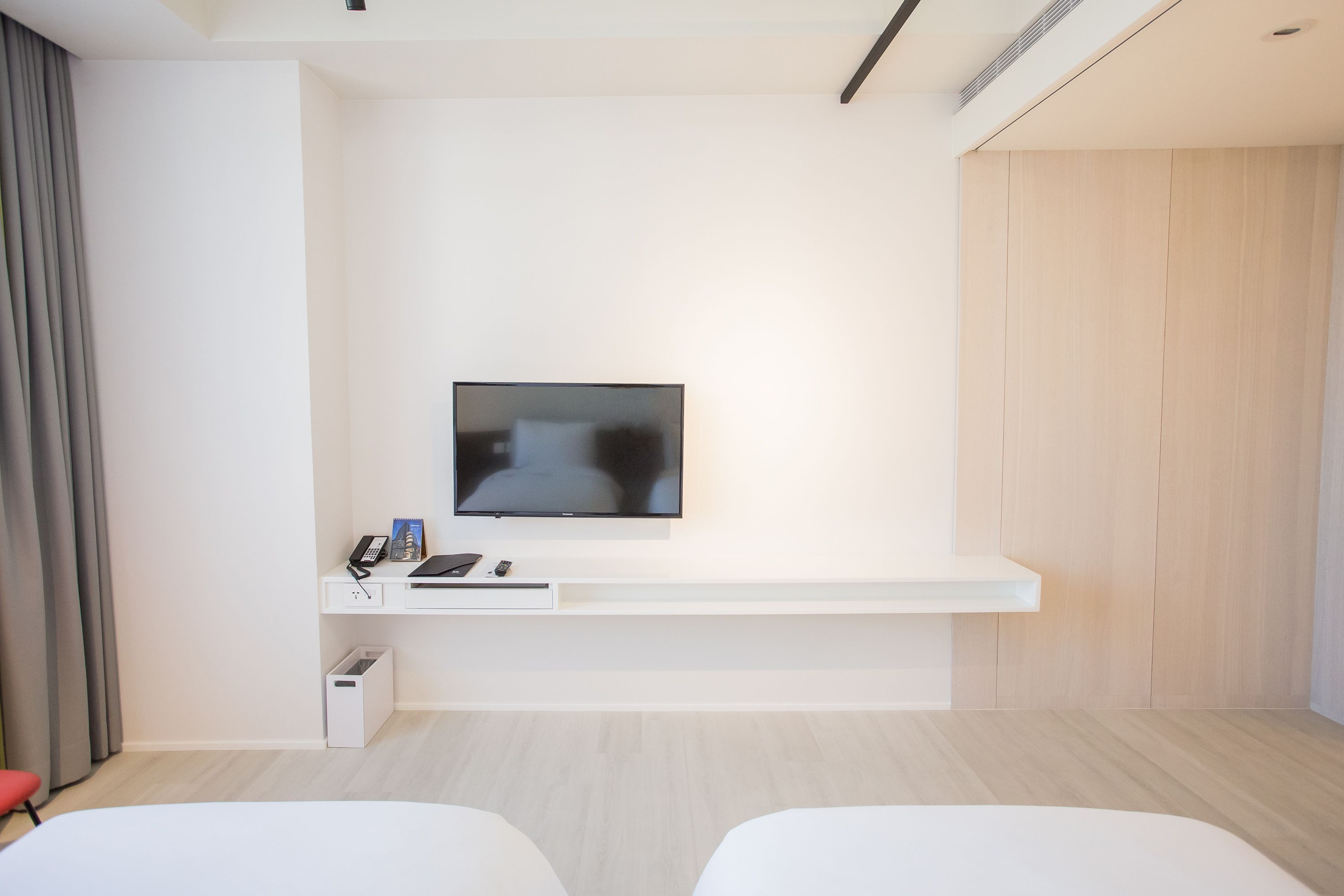 A modern hotel room with a flat-screen TV mounted on a white wall, a small desk with a phone and a laptop, and two white beds.