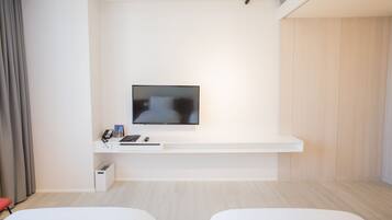 A modern hotel room with a flat-screen TV mounted on a white wall, a small desk with a phone and a laptop, and two white beds.