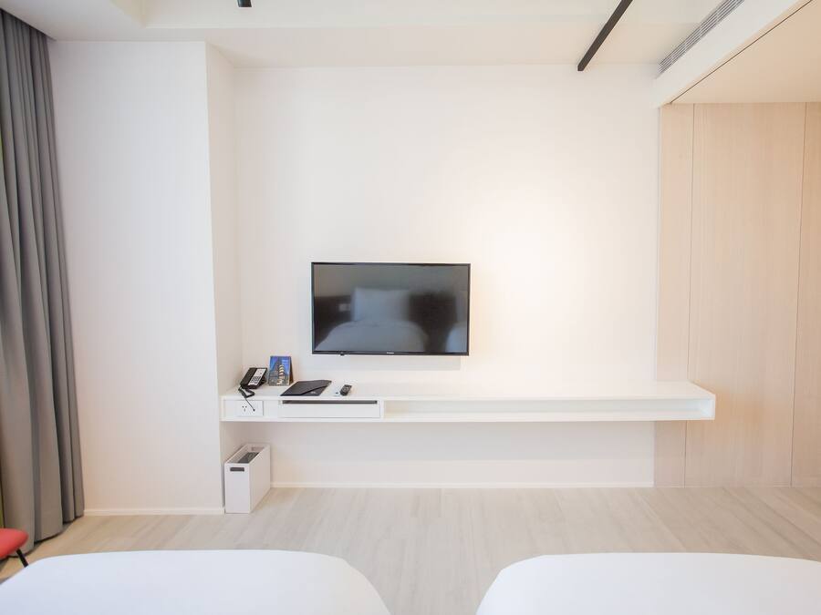 A modern hotel room with a flat-screen TV mounted on a white wall, a small desk with a phone and a laptop, and two white beds.