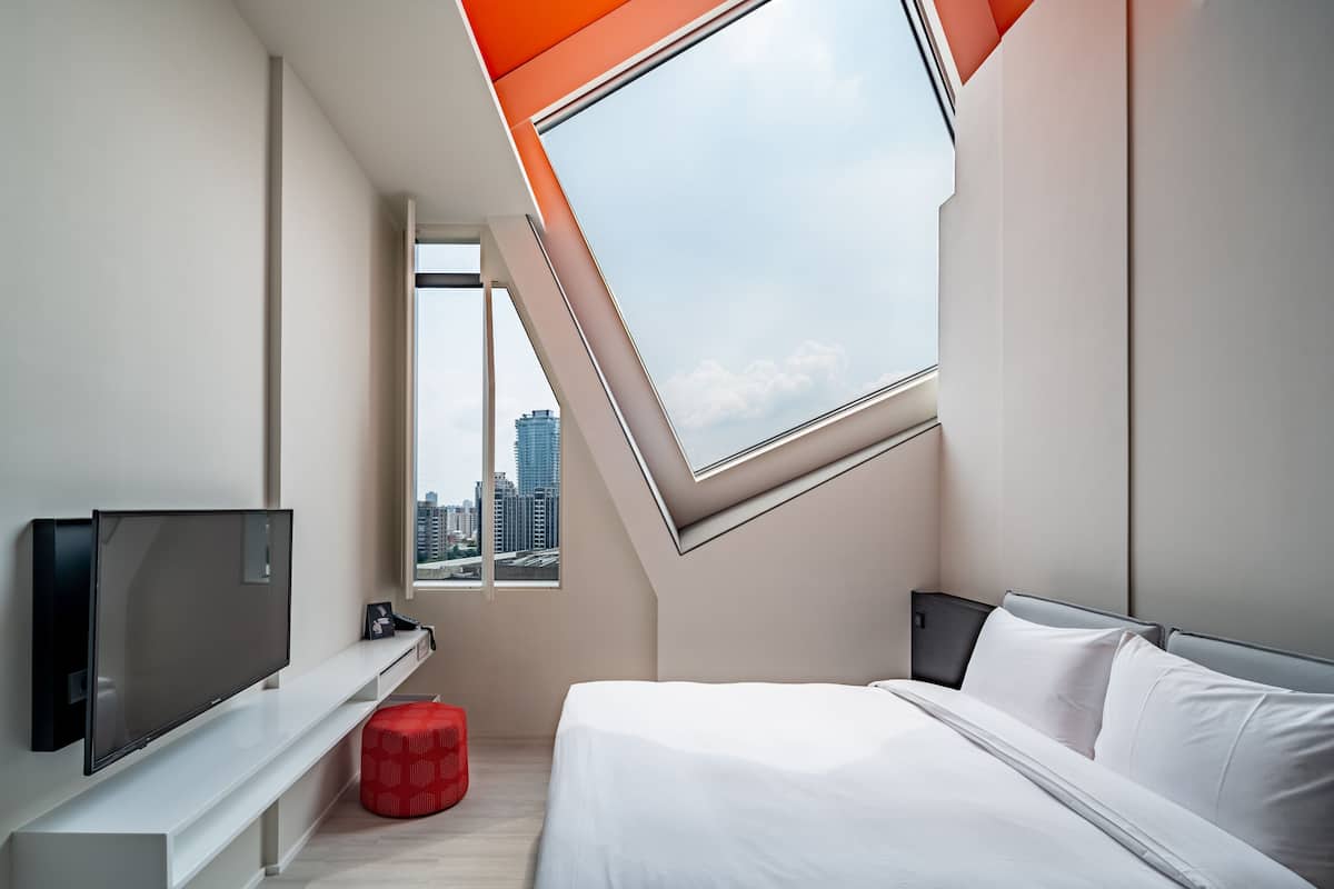 A modern bedroom with a skylight, a flat-screen TV, a bed, and a red ottoman.