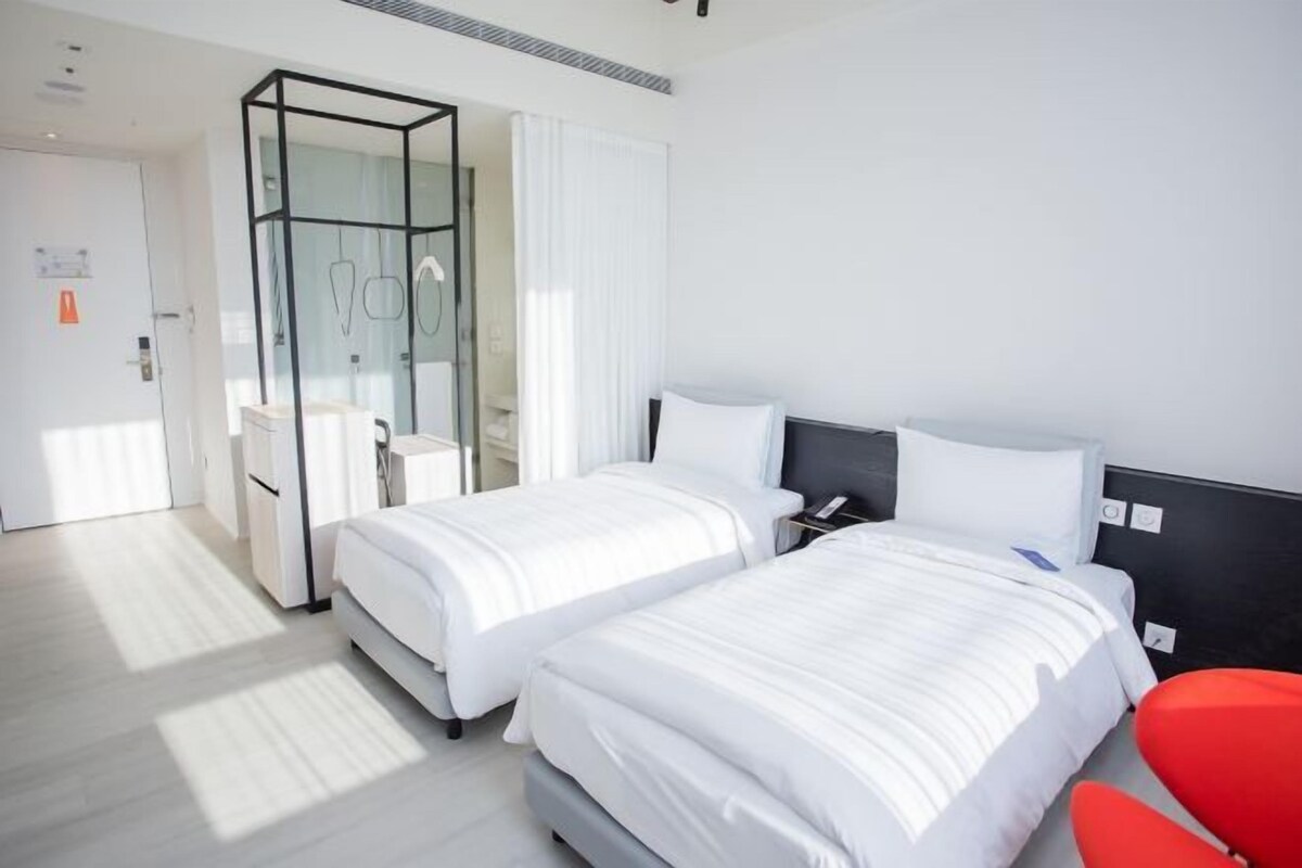 A hotel room with two beds, a red chair, and a glass partition.