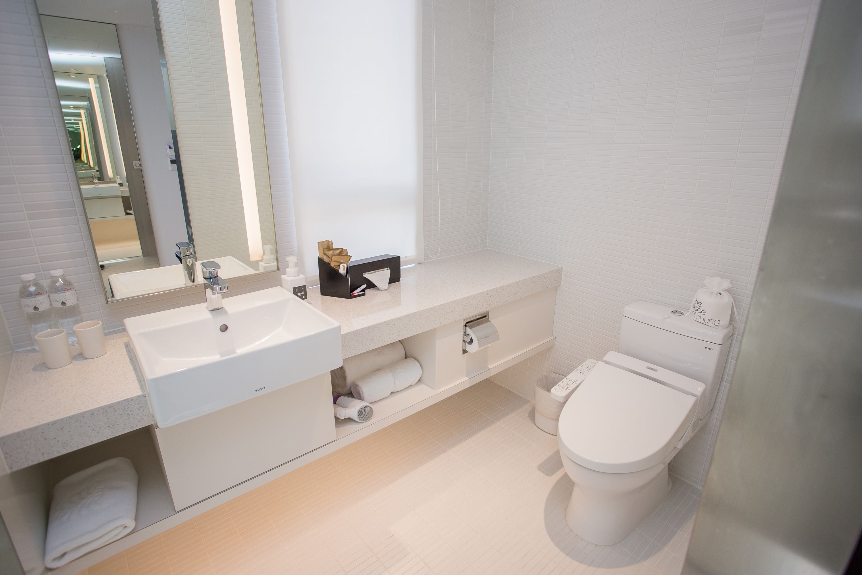 A modern bathroom with a white sink, mirror, and toilet. There are towels and toiletries on the counter.