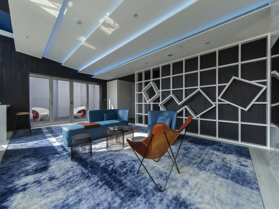 A modern living room with a blue carpet, a blue sofa, a brown leather chair, and a geometric wall design.