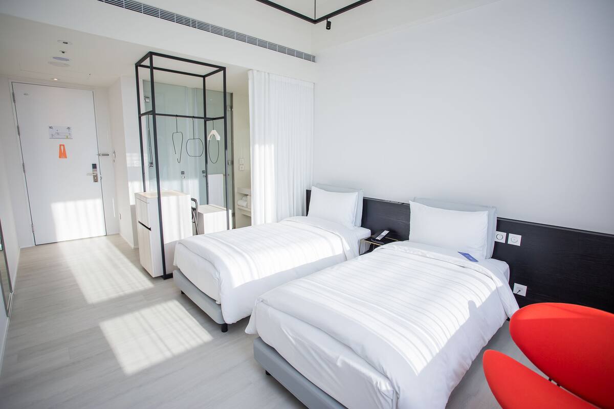 A hotel room with two beds, a red chair, and a glass shower enclosure.