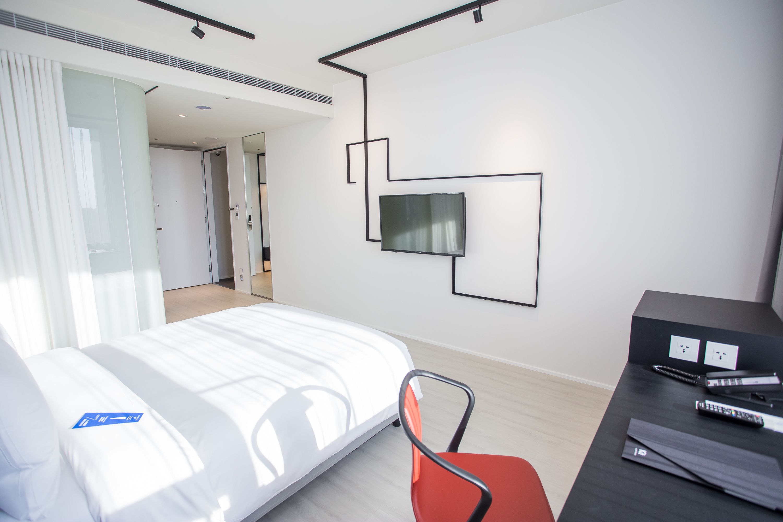 A modern hotel room with a bed, a desk with a chair, a flat-screen TV mounted on the wall, and a door leading to another room.