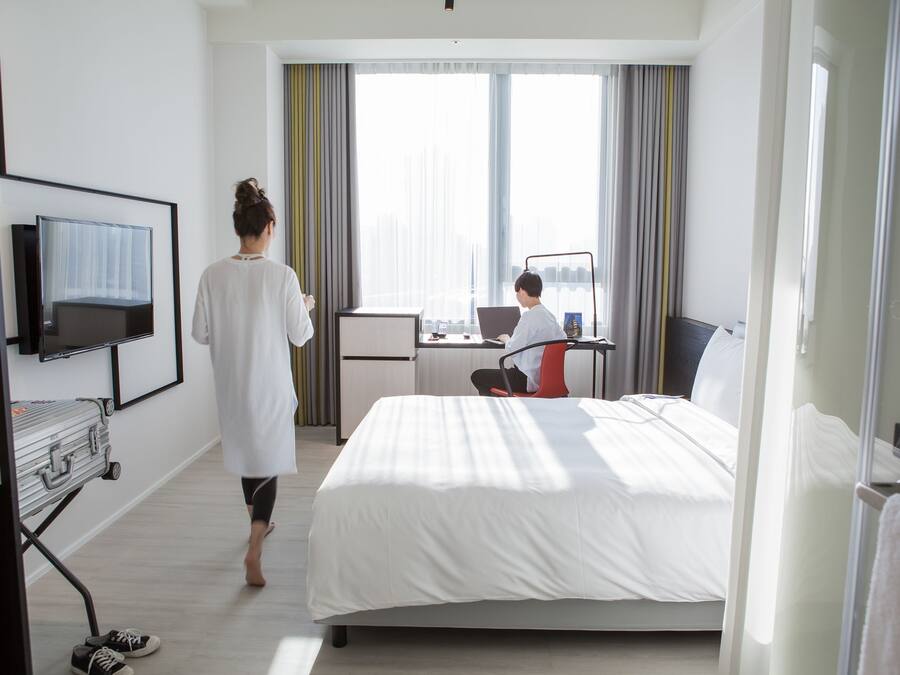 Une chambre d’hôtel avec un lit, un bureau avec un ordinateur portable, une télévision et une fenêtre avec des rideaux.