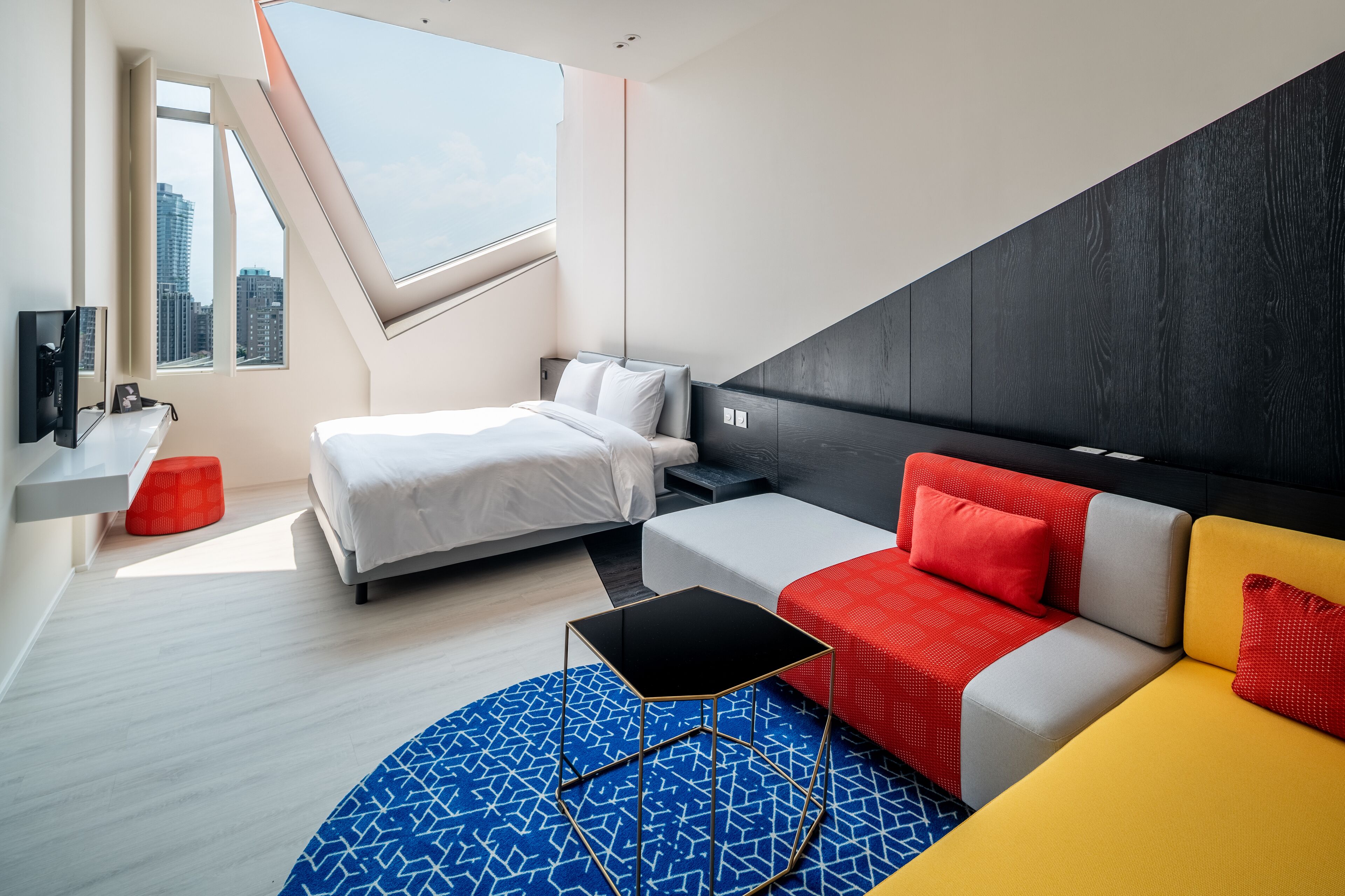 A modern hotel room with a bed, a sofa, a small table, a TV, and a window with a city view.