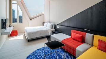 A modern hotel room with a bed, a sofa, a small table, a TV, and a window with a city view.