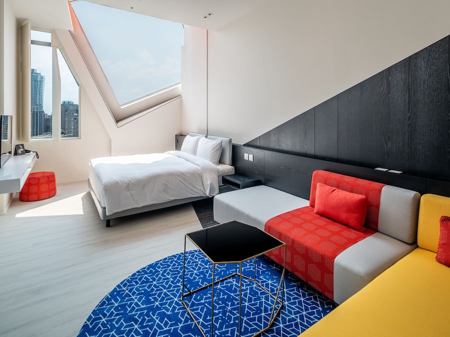 A modern hotel room with a bed, a sofa, a small table, a TV, and a window with a city view.