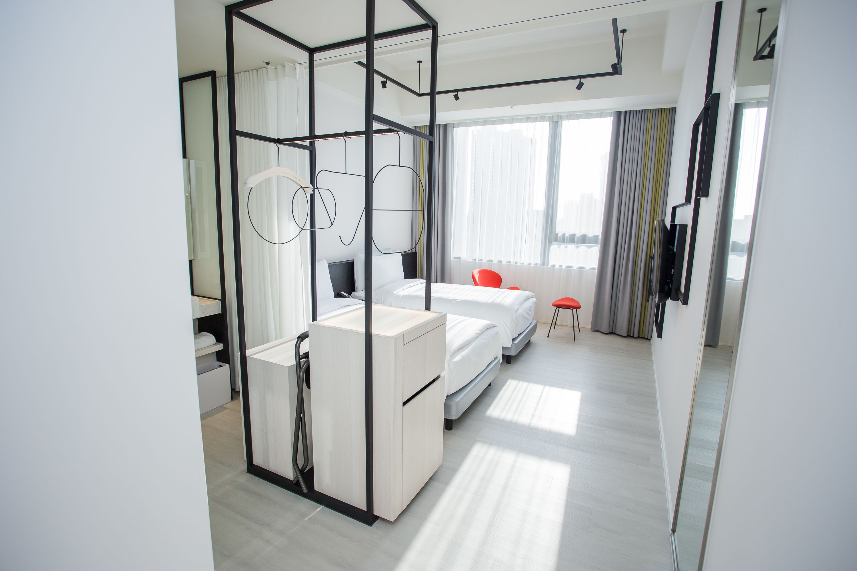 Une chambre d’hôtel moderne avec un lit, un bureau et une chaise. Il y a une grande fenêtre avec des rideaux, et un miroir sur le mur.
