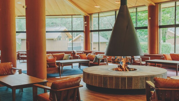 A spacious indoor area with a central fire feature, wooden furniture, and large windows offering views of the outdoors.