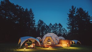 A series of dome-shaped tents with a central wooden platform and seating area, surrounded by a forested area.