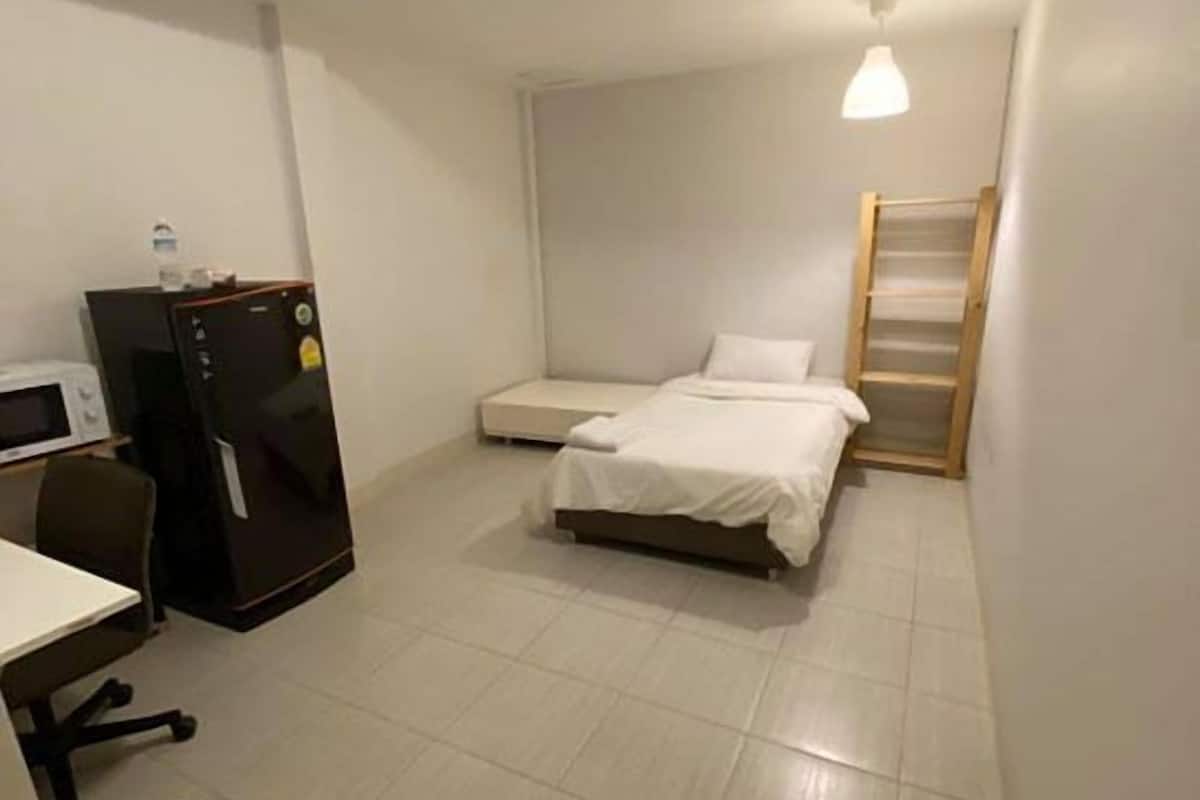 A small, clean room with a bed, a desk, a chair, a refrigerator, and a trash bin.