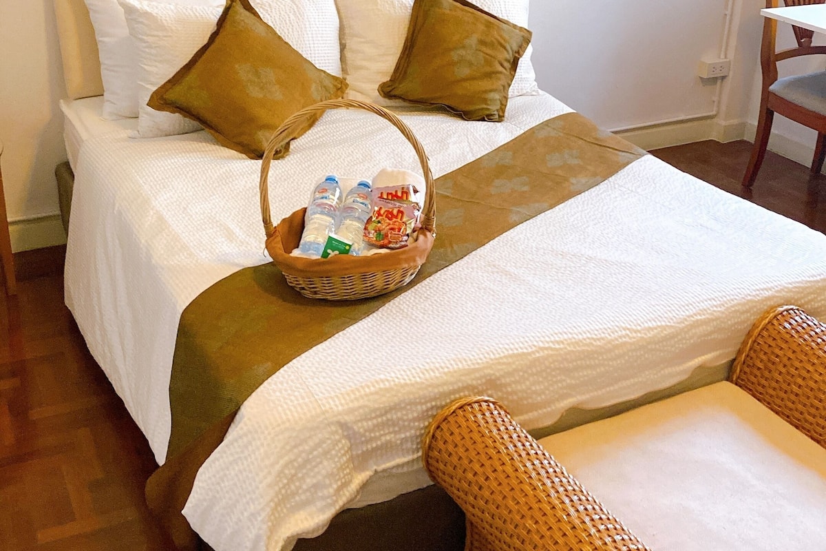 A neatly made bed with a basket of toiletries, a woven chair, and a patterned rug.