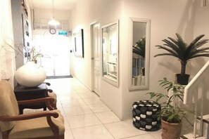 A hallway with a white tiled floor, a wooden chair, a large white vase, a potted plant, and a black and white ottoman. - 12/14 HOME Studio (Bangkok)