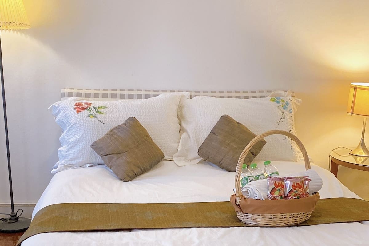 A neatly made bed with white linens, two brown pillows, a brown throw blanket, and a basket with snacks and water bottles.
