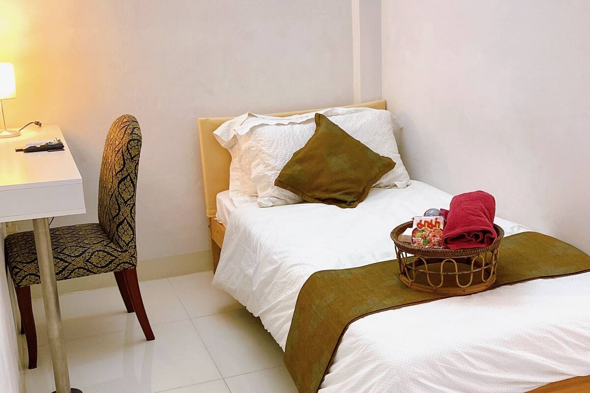 A small, neatly arranged bedroom with a single bed, a chair, a desk, and a lamp.