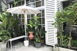 A patio area with a white umbrella, potted plants, a bench, and a white building with horizontal slats. - 12/14 HOME Studio (Bangkok)