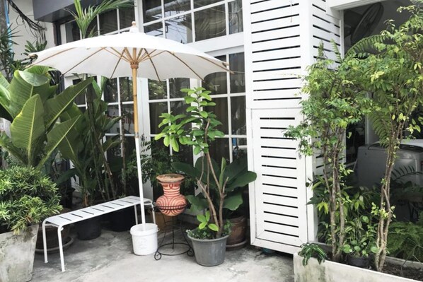 Un patio con sombrilla blanca, plantas en macetas, un banco y un edificio blanco con listones horizontales.