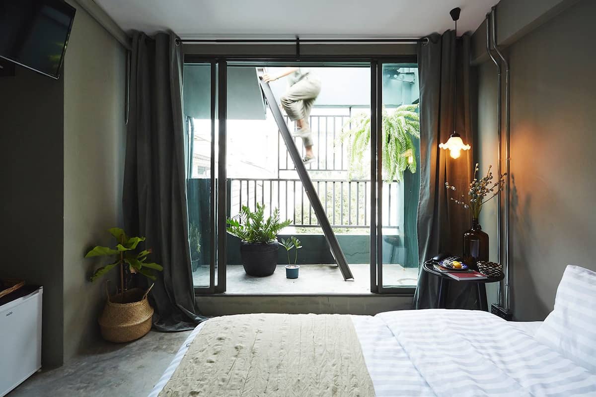 A bedroom with a large bed, a window with a view of a balcony, a potted plant, and a bedside table with a lamp.