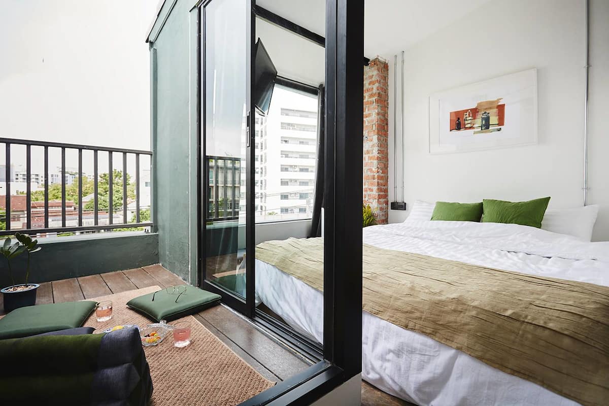 A modern bedroom with a large bed, a balcony with a view of buildings, and a small seating area with cushions and a plant.