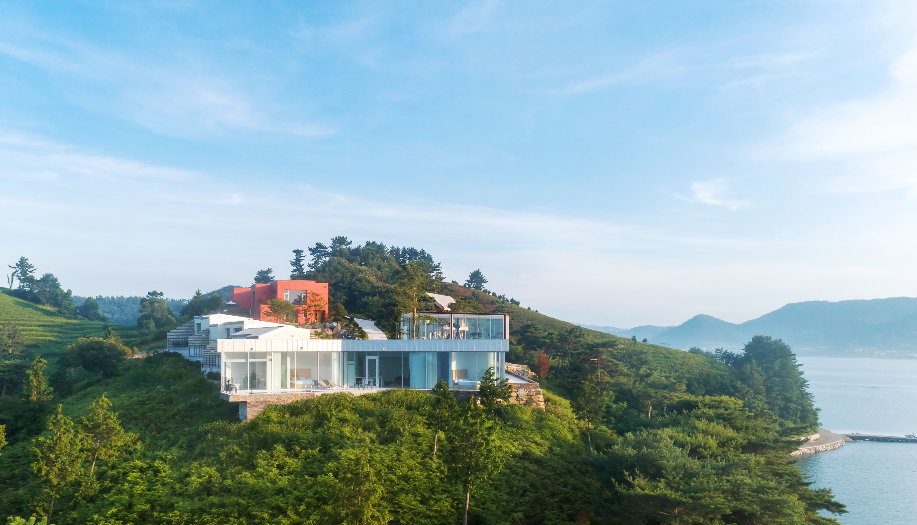 A modern house with a glass facade, situated on a hillside overlooking a body of water.