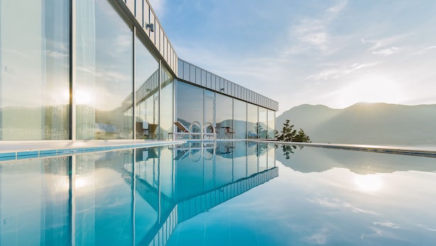 A modern infinity pool with clear water reflecting the sky and mountains, surrounded by a glass building with outdoor seating and a view of the landscape. - Bayclub (Namhae)