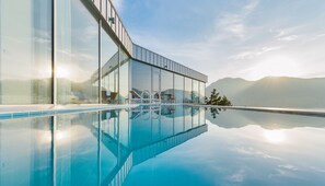 Una piscina infinita moderna con agua cristalina que refleja el cielo y montañas, rodeada por un edificio de vidrio con asientos al aire libre y vistas al paisaje.