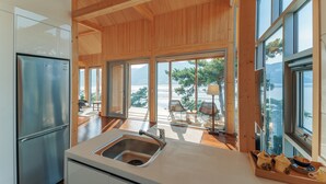 A modern kitchen with a stainless steel refrigerator, a sink, and wooden flooring. Large windows offer views of a balcony with outdoor furniture. - Bayclub (Namhae)