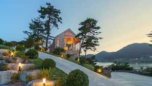 A modern house with a large glass window, surrounded by trees and a view of mountains. - Bayclub (Namhae)