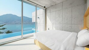 A modern bedroom with a large window, a bed, and a view of a body of water and distant landscape. - Bayclub (Namhae)