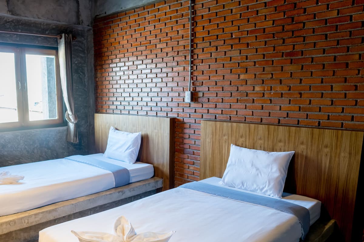 Two single beds with wooden headboards and white bedding, set against a brick wall with a window to the left.