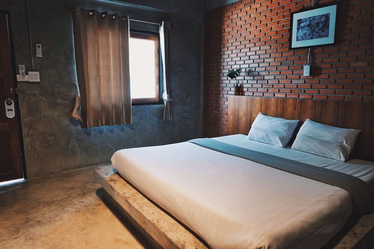 A bedroom with a bed, a window with curtains, a brick wall, and a picture on the wall.