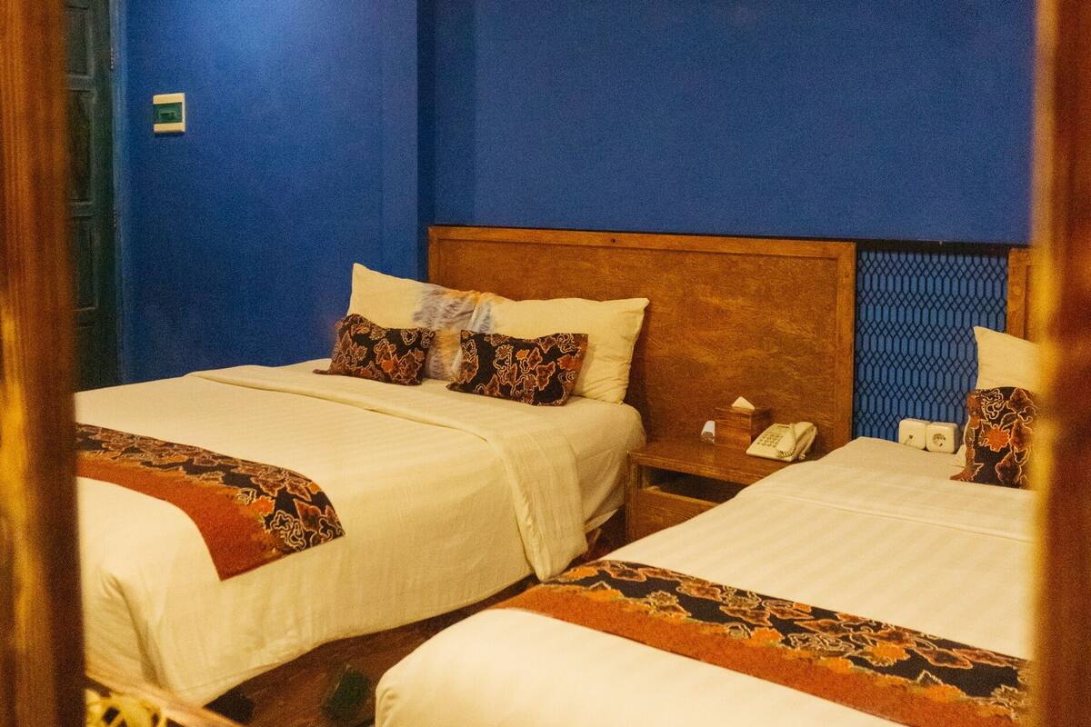 A hotel room with two beds, each with white linens and patterned pillows. The room has blue walls and a wooden headboard.